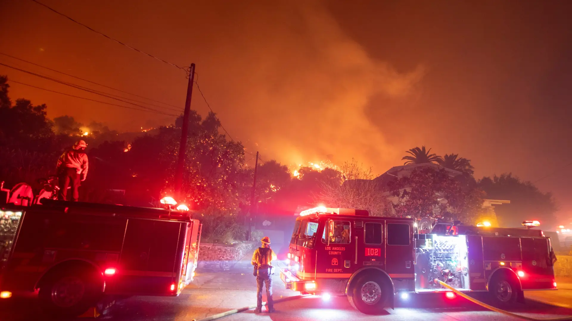 Firefighters and fire trucks responding to a wildfire near homes at night, highlighting the urgency of fire watch security in wildfire prevention.