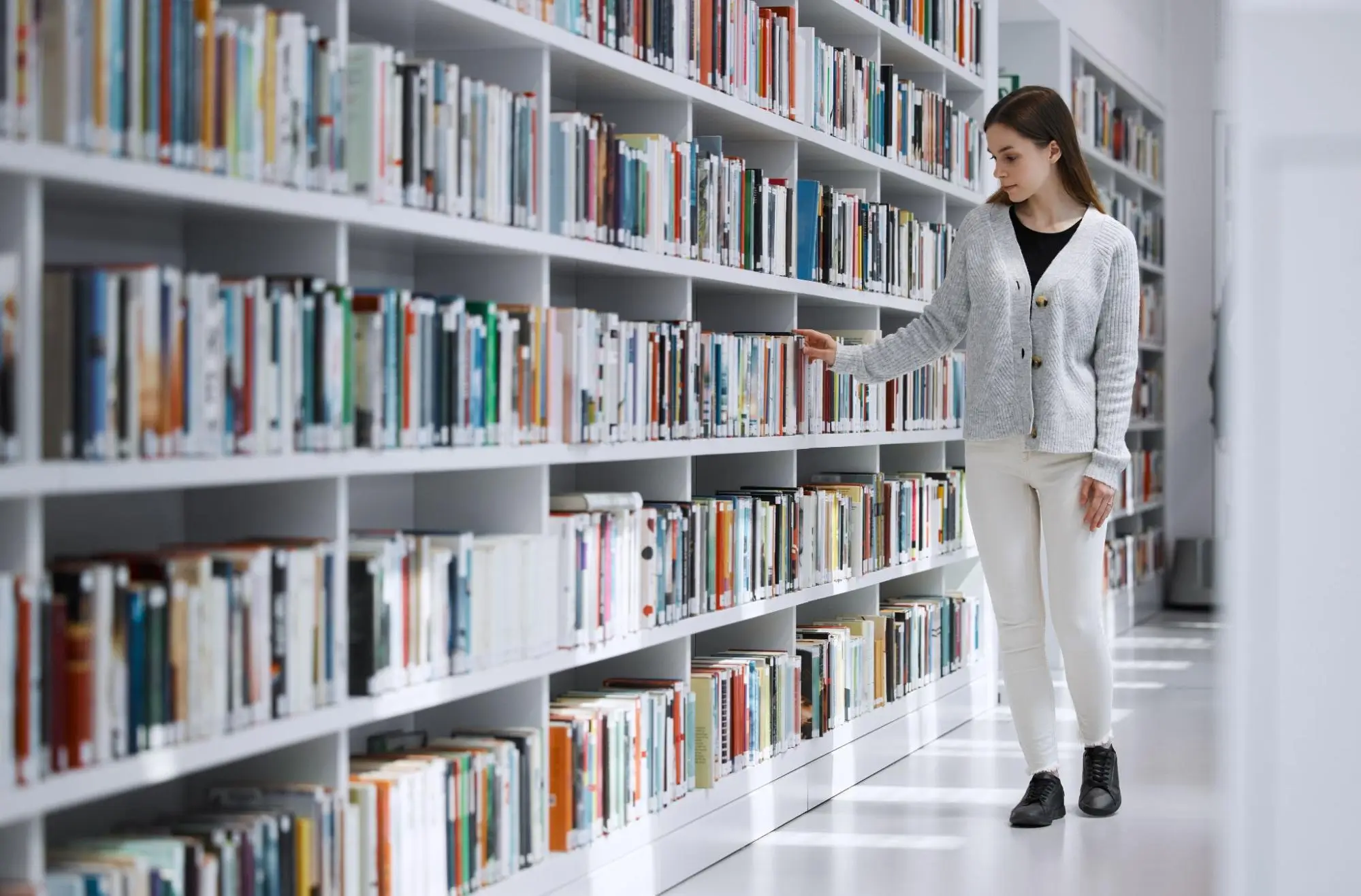 Library Security Woman browsing books along a bright white library shelf, reaching for a title in a clean, well-organized space that reflects safe, well-managed library security and access